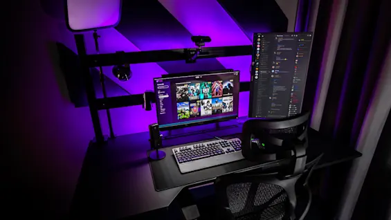Et moderne sort skrivebord med to skjermer; en viser et spillbibliotek med titler som Apex Legends, Among Us, Minecraft. Et lilla LED-lys lyser opp den ergonomiske stolen og omgivelsene.