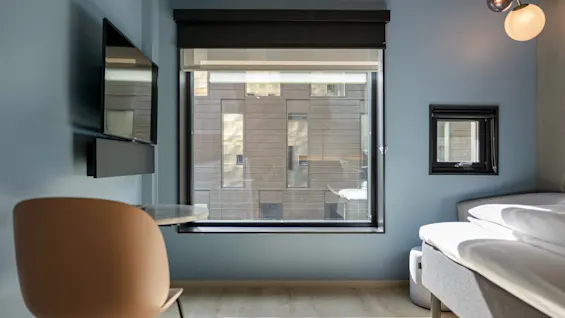 Et lyst og moderne hotellrom badet i naturlig lys, med en komfortabel seng, et vindu med byutsikt og et lite sitteområde.