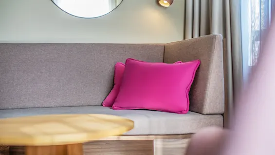 A gray sofa holds two bright pink pillows, positioned in a modern living room with a wooden table, wall mirror, and soft lighting from a wall-mounted lamp beside a window.