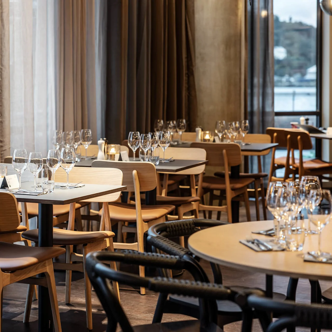 Borde dækket med vinglas og servietter står i en elegant restaurantspisestue med lyse træstole, omgivet af store vinduer og bløde, flydende gardiner.