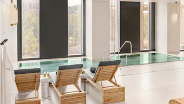 Sunchairs with cushions face an indoor swimming pool, adjacent to large windows. A small table holds glasses of champagne. A robe hangs nearby with text "elements spa."