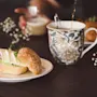 A sandwich with cheese and greens rests on a plate beside a floral-patterned mug being filled with tea; a person holds a cup in the background on a wooden table.