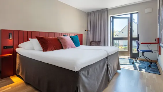 Two twin beds with white linens and colorful pillows in a neat, modern hotel room with window and work desk.