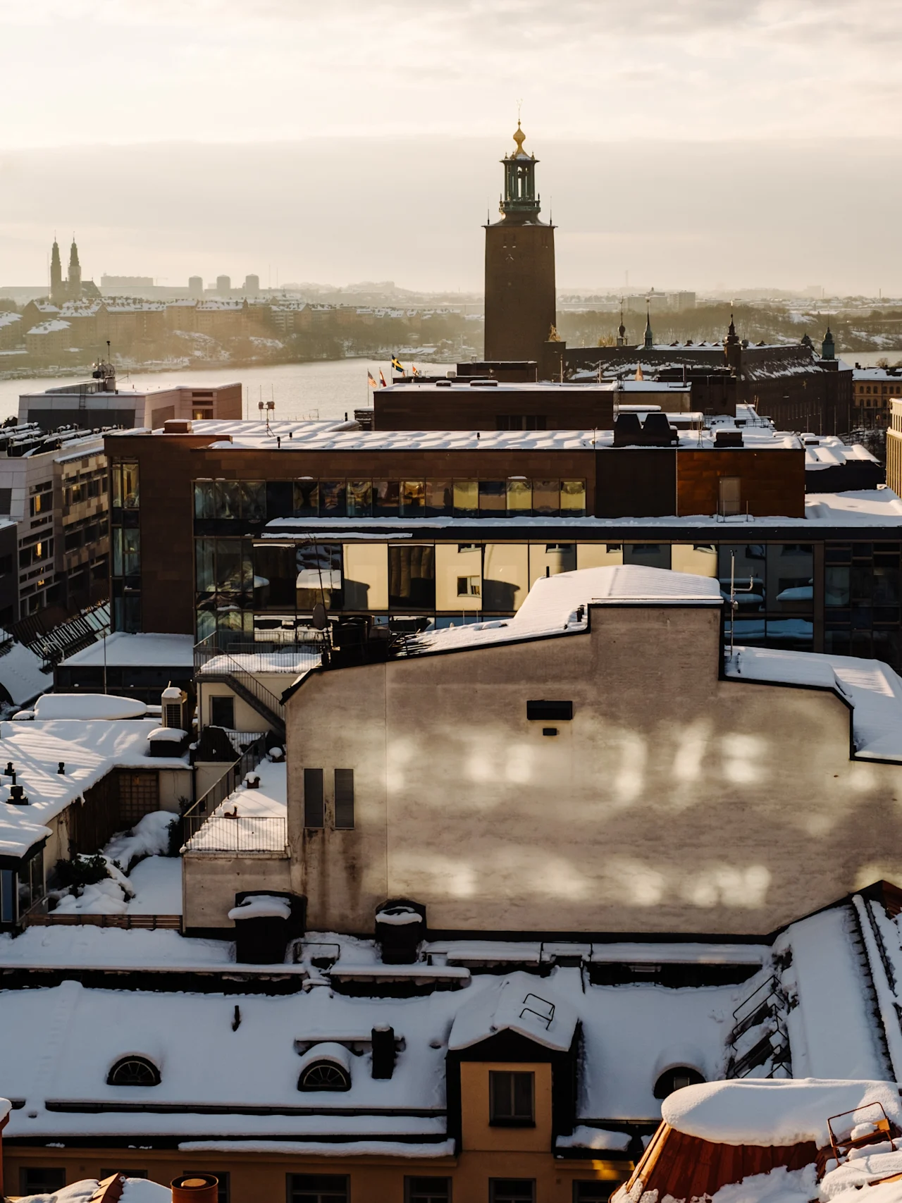 Swedish: Tak täckta av snö omger en hög byggnad med ett torn. Stadsbilden ses vid solnedgången, vilket kastar ett varmt ljus över stadslandskapet.