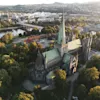Luftfoto av Nidarosdomen i Trondheim omkranset av grønne trær i mykt sollys.