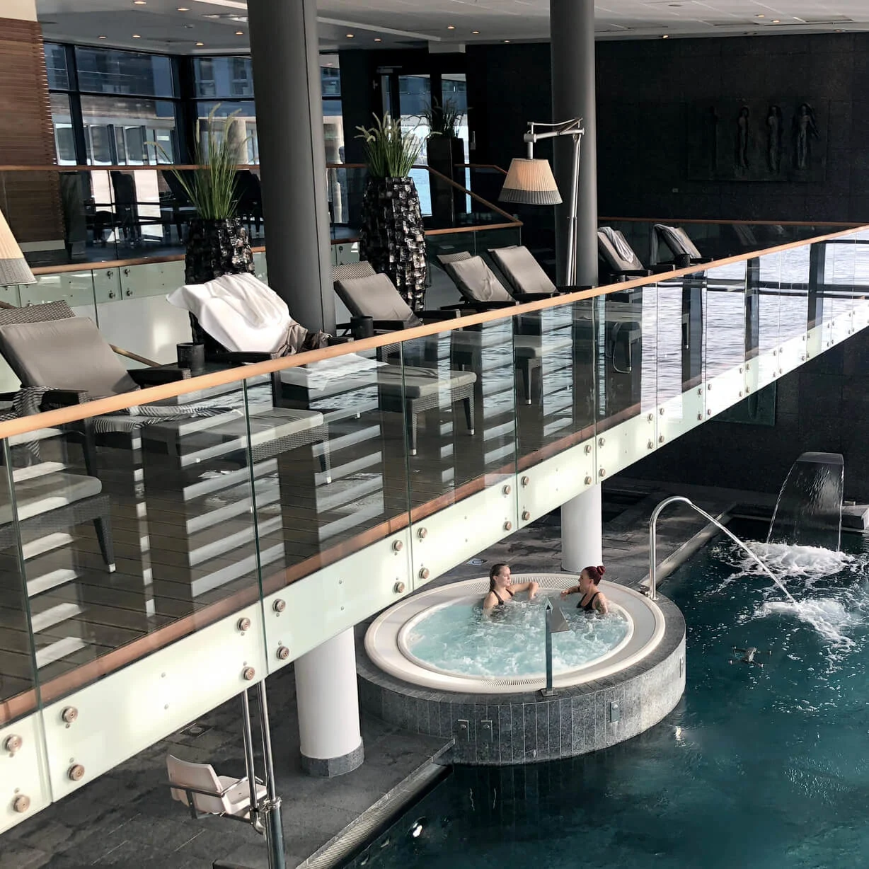 Et indendørs spa med en pool, hvor to personer slapper af, med udsigt til havet gennem store vinduer.
