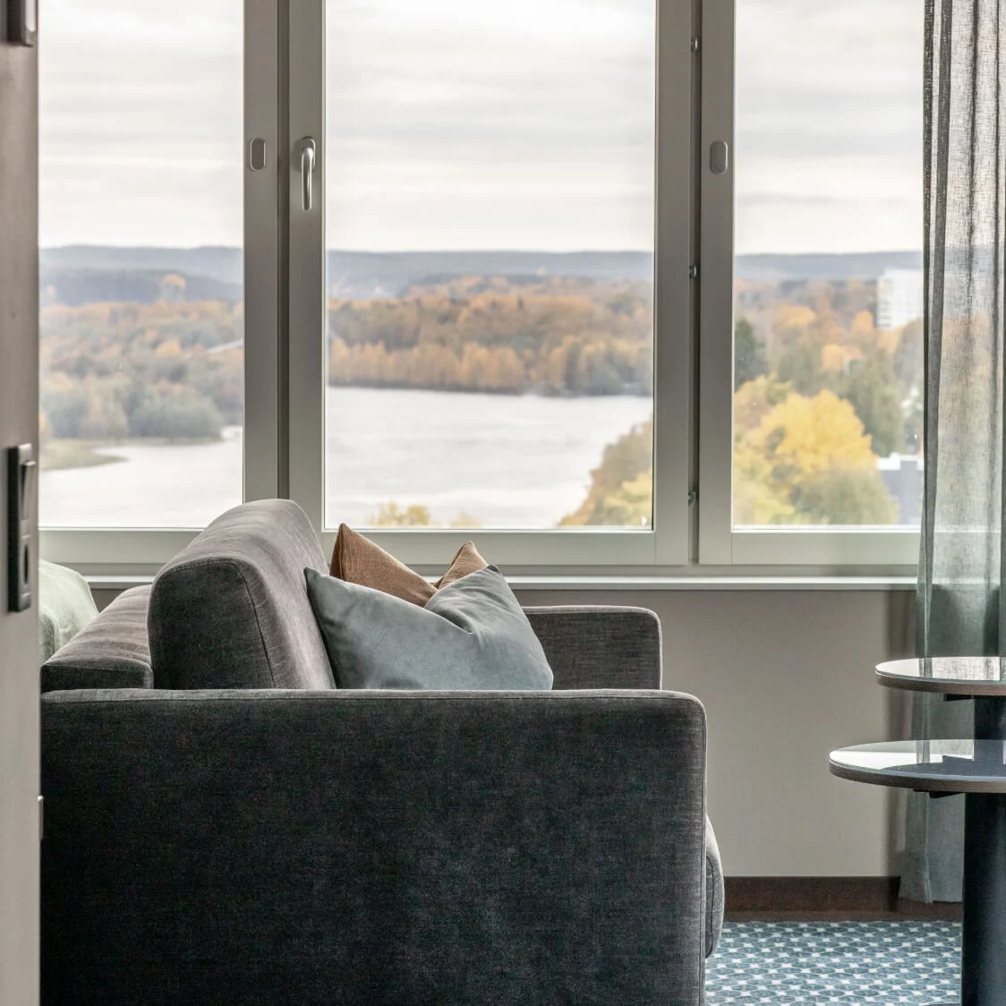 Grå sofa vender mod stort vindue med udsigt over efterårstræer ved en sø; runde borde ved siden af sofa; rum med blå gardiner og mønstret tæppe.