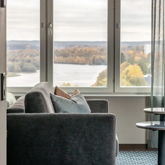 Grå soffa mot stort fönster med utsikt över höstträd vid en sjö; runda bord bredvid soffa; rum med blå gardiner och mönstrad matta.