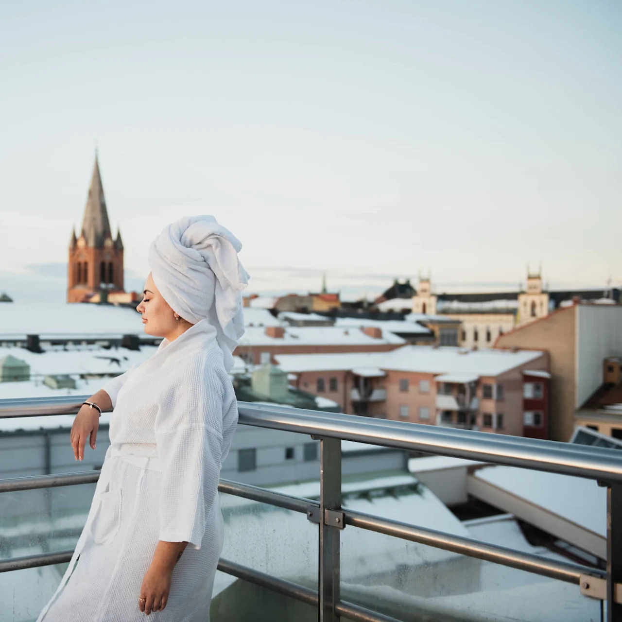 A person in a bathrobe and towel relaxes on a balcony above snow-covered rooftops, with a church spire visible in the background.