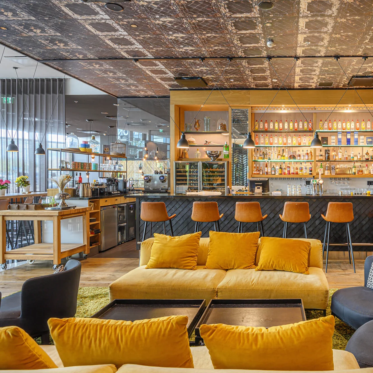 A bar with amber stools and a black counter displays bottles on wooden shelves. Yellow couches and tables, surrounded by modern decor, create a cozy lounge atmosphere.