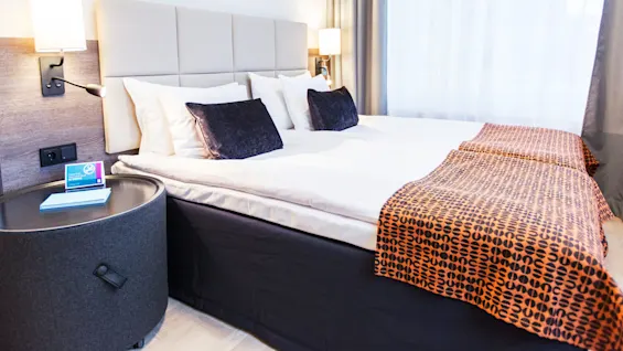 En säng bäddad med vita lakan och ett mönstrat orange överkast flankeras av lampor på sängbord i ett ljust hotellrum. En skylt på bordet lyder "Quality time with high speed internet".