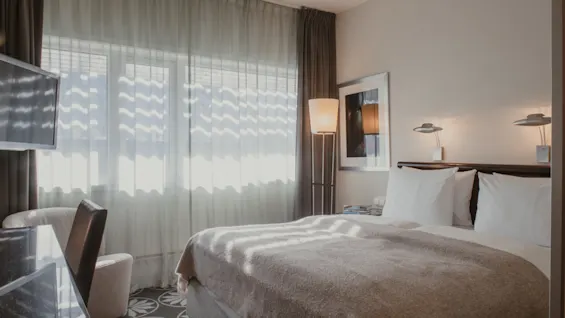 Ett ljust hotellrum med en bekväm säng, ett fönster med skira gardiner och varmt solljus som strömmar in.