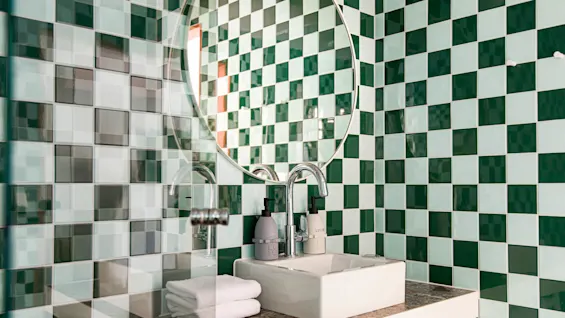 En hvid vask holder sæbe- og lotiondispensere på en marmor disk, omgivet af grønne ternede fliser og reflekteres i et rundt spejl.