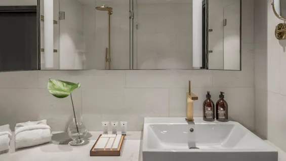 Et moderne baderom har en hvit vask med gullkran, ved siden av toalettartikler, en glassvase med et grønt blad, brettede håndklær og en dusj i bakgrunnen.