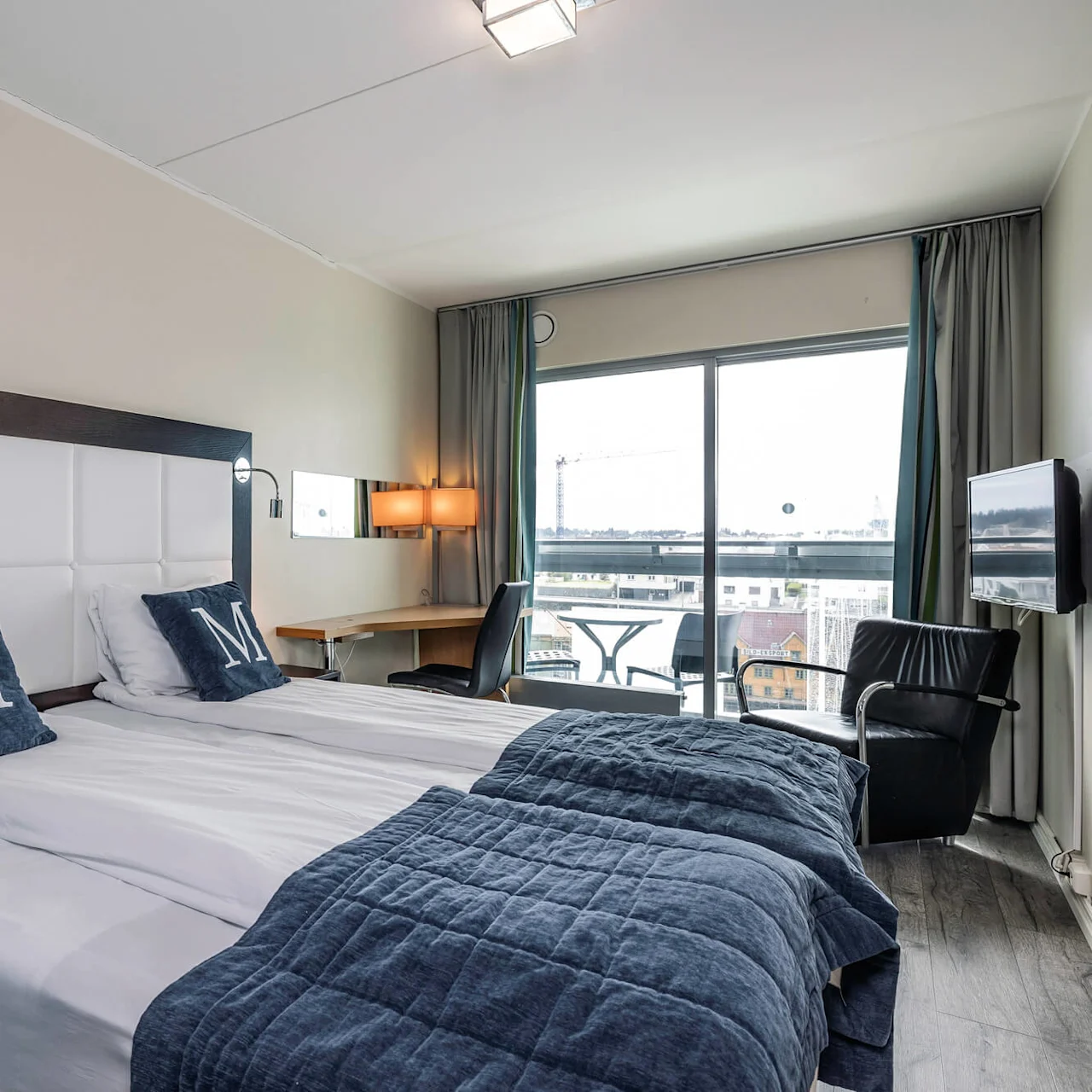 Ett hotellrum med två enkelsängar, varje med kuddar med "M" på, har utsikt över stadsmiljön genom stora fönster. Ett skrivbord, en stol och en fåtölj förbättrar dess funktionalitet.