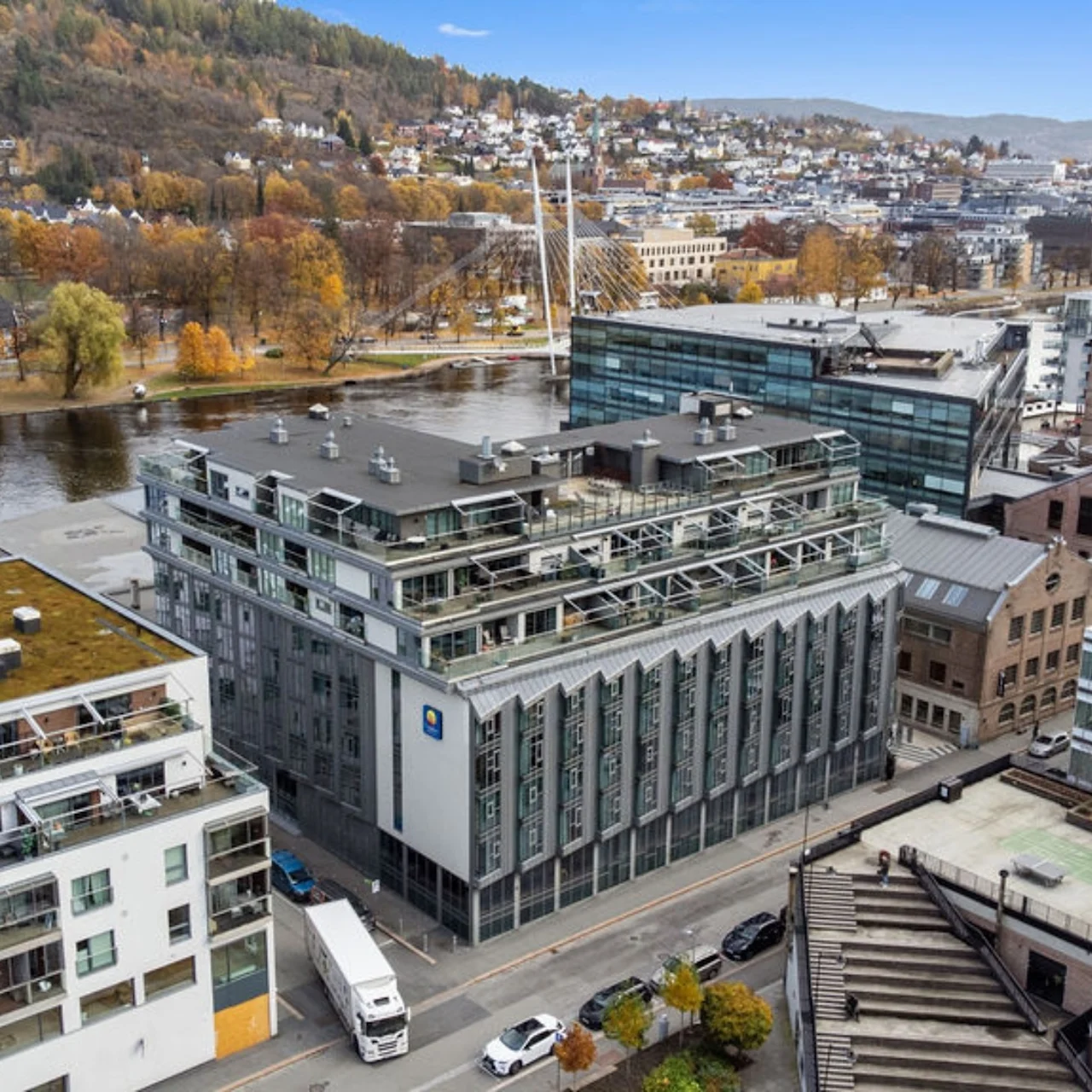 A large, modern office building stands by a river, surrounded by other urban structures and trees. Several vehicles are parked on the adjacent street. Autumn foliage colors the hillside in the background.