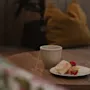 En kopp te står på ett träbord bredvid en tallrik med kaka och jordgubbar, i ett mysigt rum med kuddar.