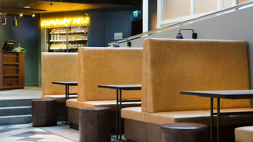 High-backed tan sofas and round stools furnish a modern cafe space; a neon sign reads "COMFORT DELI".