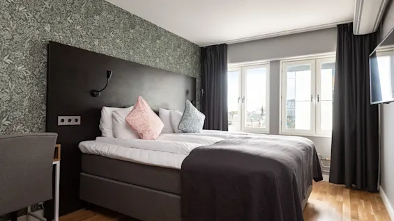 A neatly made bed in a stylish bedroom with a dark headboard, patterned wallpaper, and bright window view.