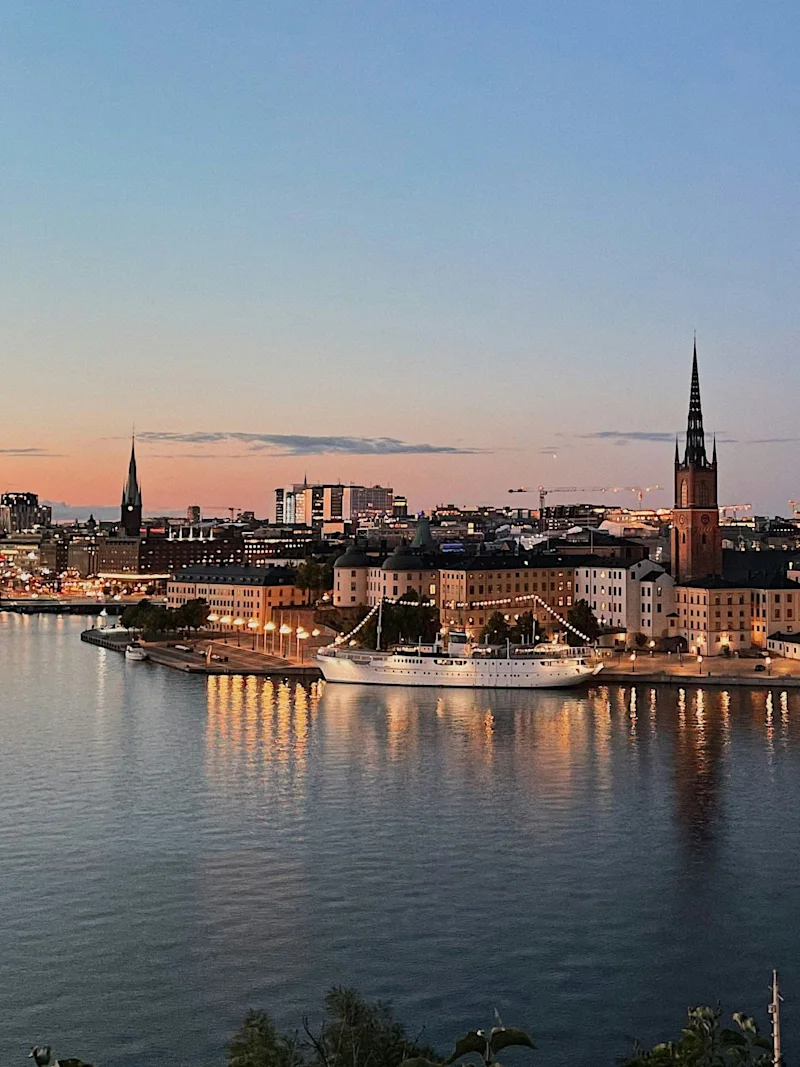 City skyline at dusk with illuminated buildings reflecting on the water, two prominent spires rise above the cityscape.