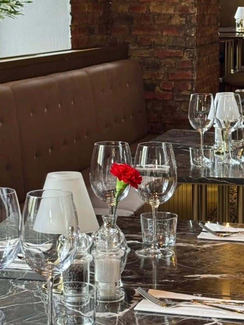 Elegant matsalsbord, prydd med tomma vinglas och en röd blomma, beläget i en restaurang med bruna lädersäten och tegelväggar.