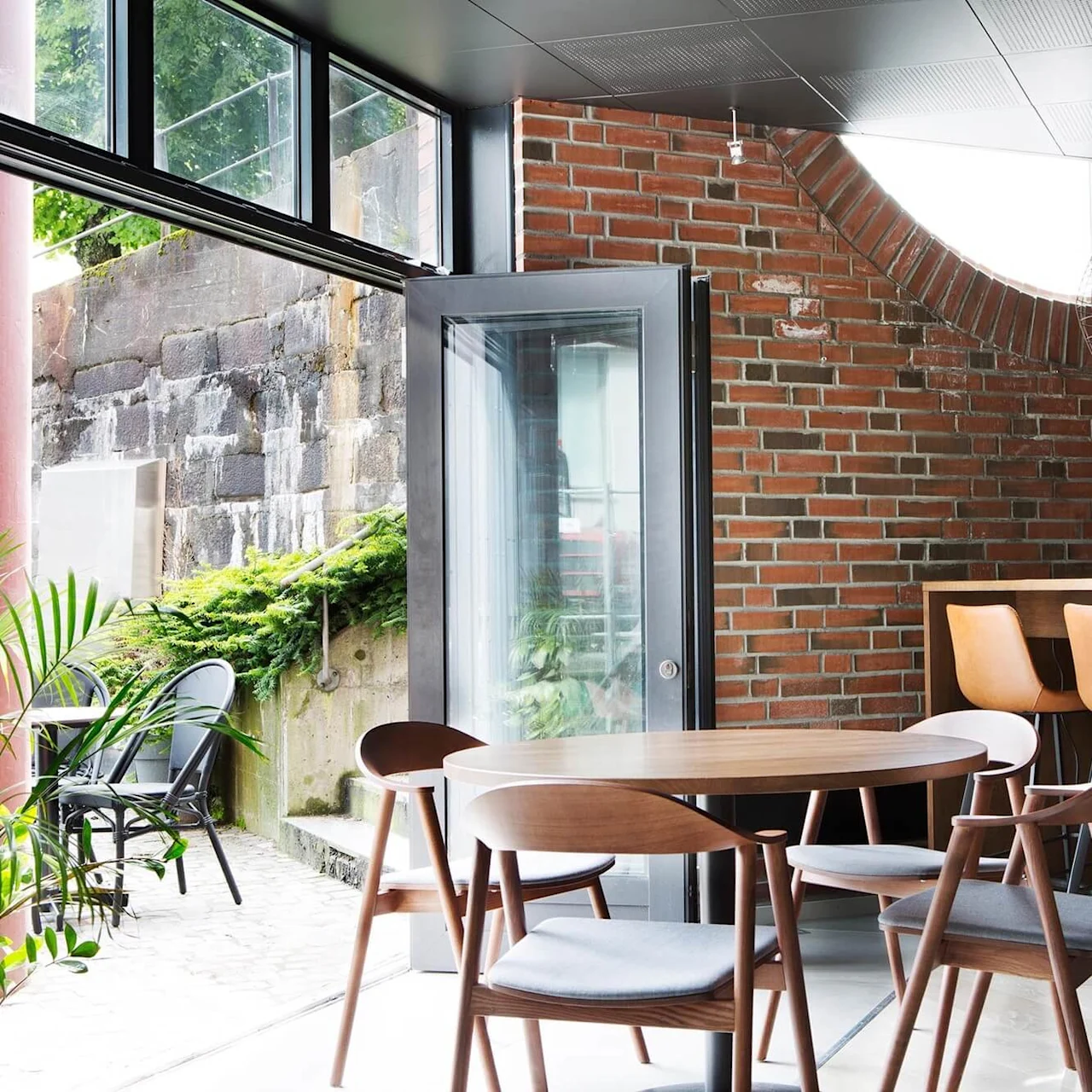 Rundt træbord omgivet af stole står i en moderne café med murstensvægge og store vinduer, ved siden af en åben dør, der fører til en udendørs terrasse med grønne planter.