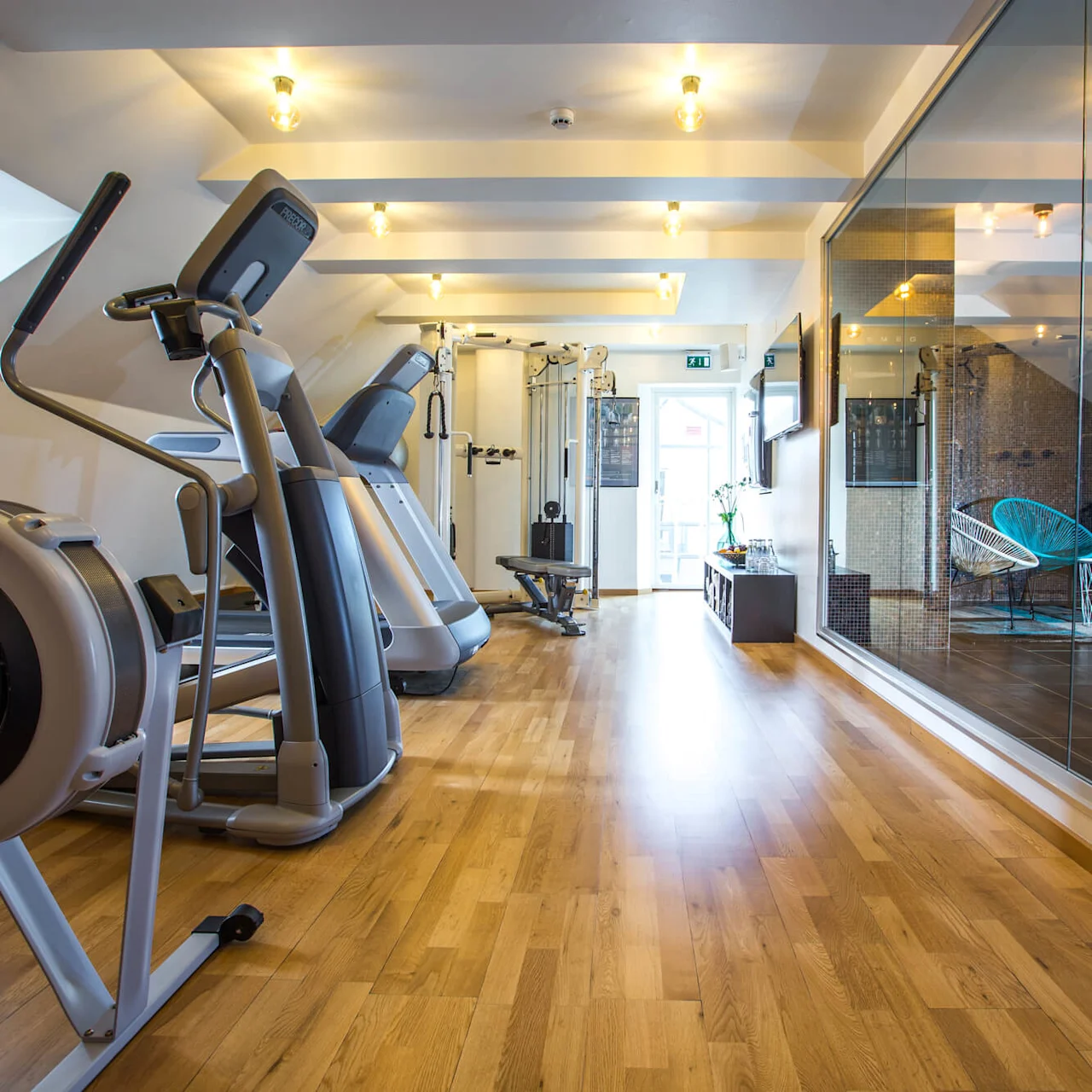 Exercise equipment in an upscale, well-lit gym with hardwood floors, mirrored walls, and a cozy seating area visible through a glass partition.