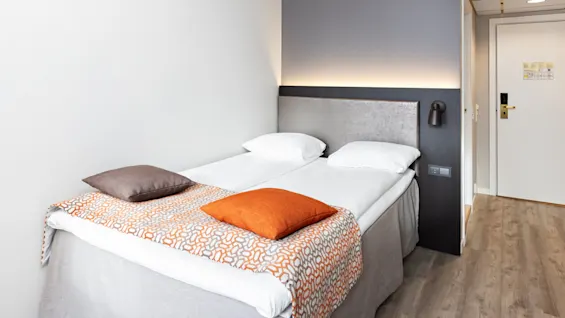 En pænt redt seng med to hvide puder, en orange-mønstret løber og farverige puder. Den står ved siden af en grå polstret sengegavl i et hotelværelse med trægulv og en lukket dør.