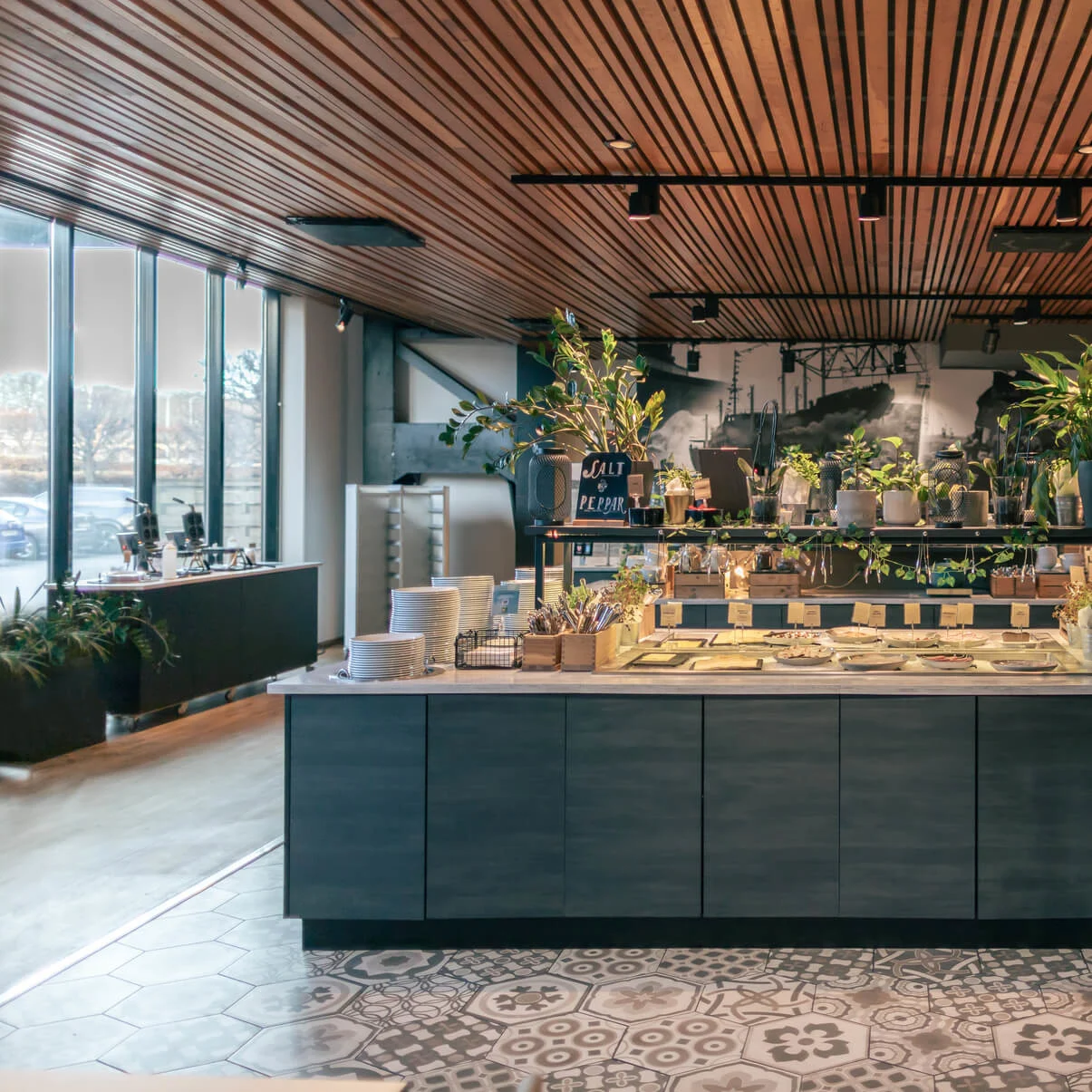 A buffet counter displays various dishes with plants as decor. Behind, there are windows bringing in natural light. A small chalkboard reads "SALT & PEBER."