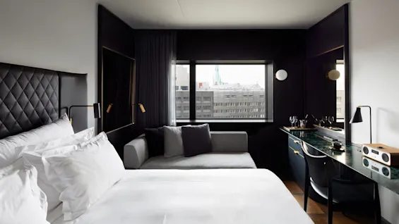 Et luksuriøst hotellrom har en stor seng med stoppet hodegjerde, en grå sofa ved et vindu med utsikt over byens bygninger og et stilig skrivebord med høyttaler og glass.
