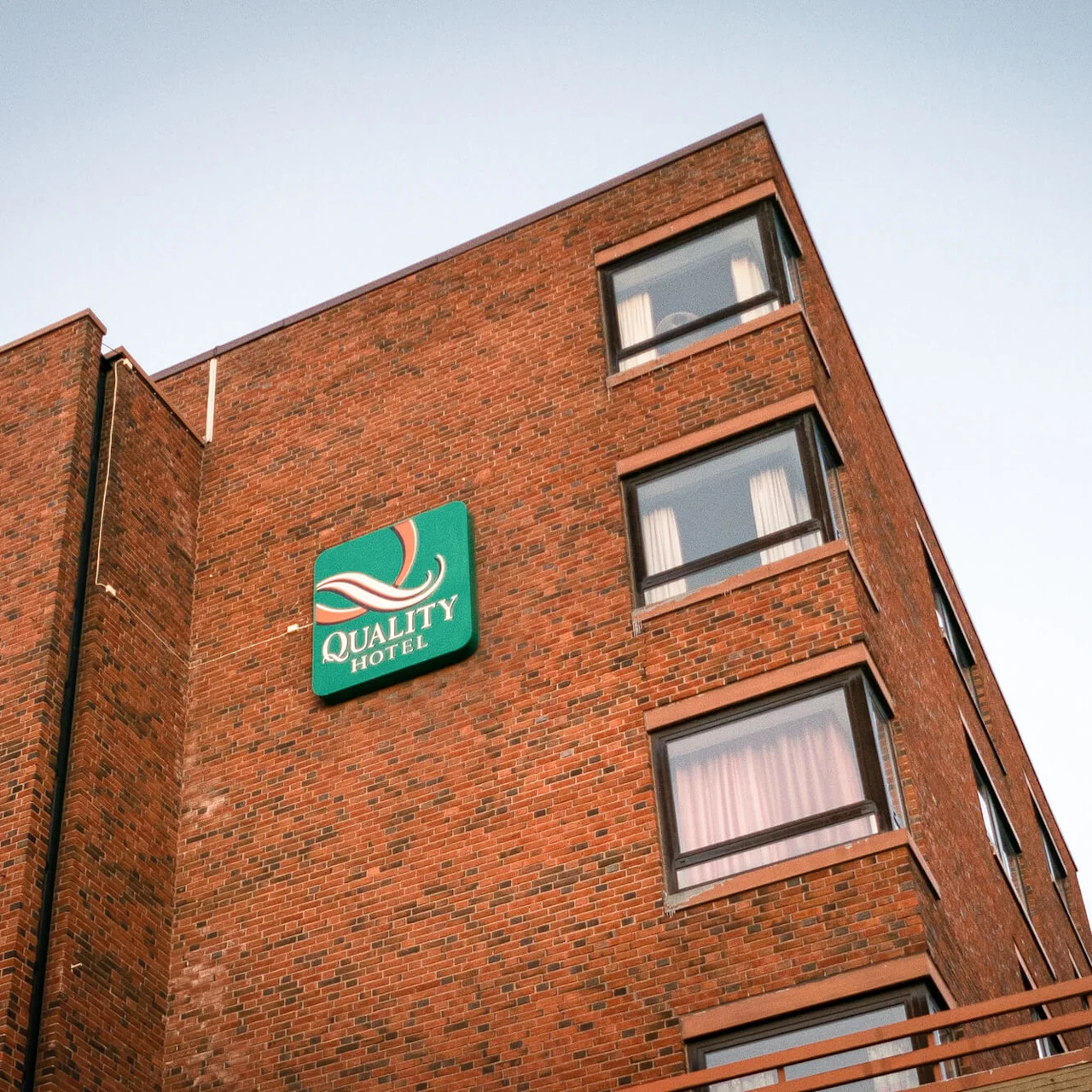 A brick building displays the "QUALITY HOTEL" sign in large white letters on a green background, with multiple windows visible against a clear sky.