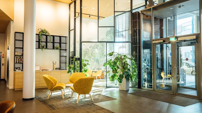 Kirkas lounge, jossa keltaisia tuoleja matalan pöydän ympärillä, lähellä suuria lasiovia. Sisäkasvit koristavat tilaa tuoden luonnollista tunnelmaa. Puu- ja metalliyksityiskohdat täydentävät modernin muotoilun.
