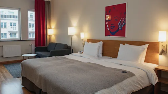 A neatly made double bed is in a cozy hotel room with modern lamps and a gray sofa. A bright red artwork is hung above the bed. Two large windows overlook neighboring buildings.