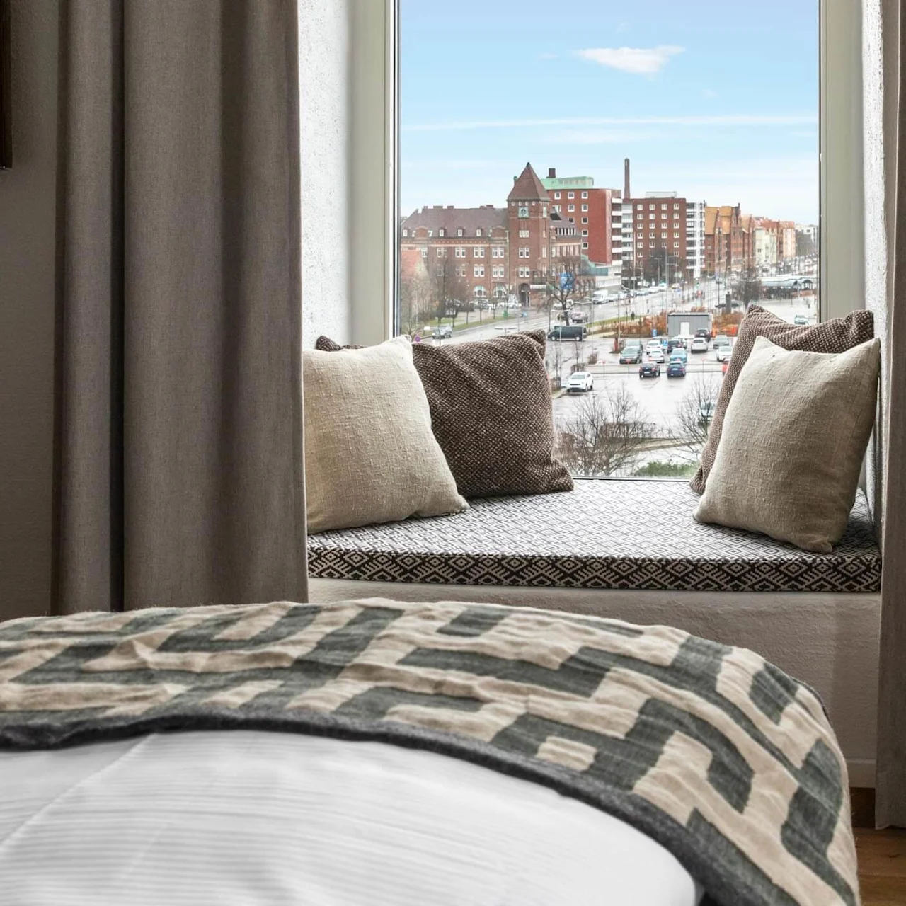 A bed foregrounding a window view of an urban landscape, with patterns and pillows enhancing interior comfort.
