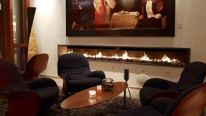 Four dark armchairs encircle a wooden coffee table, adorned with a candle and small vase, in a cozy room with a wall-mounted gas fireplace and an abstract painting above.