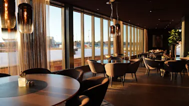 A modern restaurant or lounge with large windows overlooking a snowy landscape at sunset, featuring stylish tables and chairs.