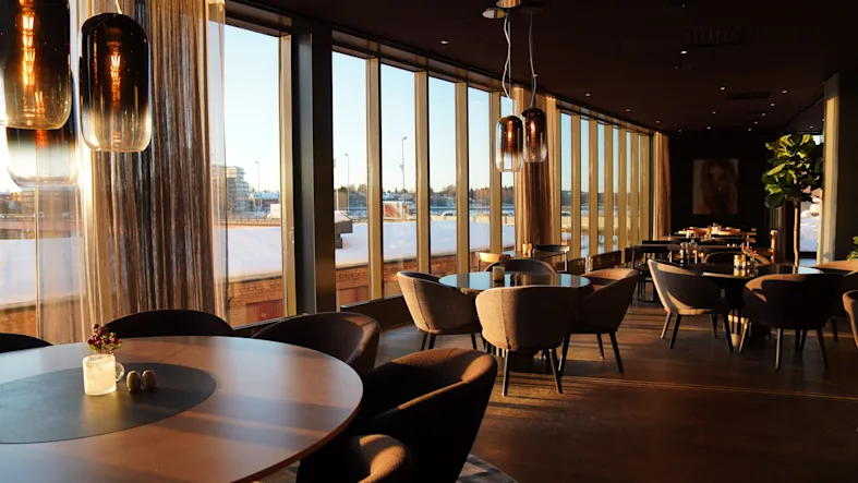 A modern restaurant or lounge with large windows overlooking a snowy landscape at sunset, featuring stylish tables and chairs.