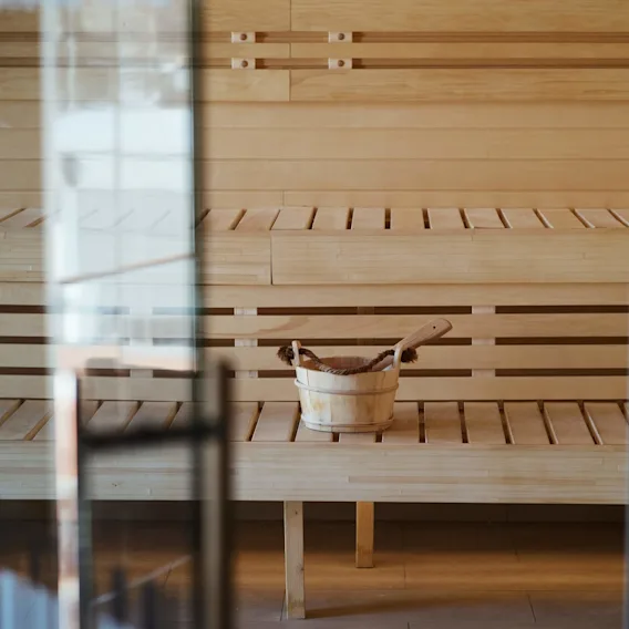 A wooden sauna bucket with a ladle rests on a bench inside a wooden sauna, viewed through a glass door.