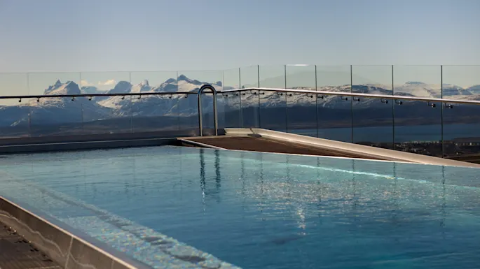 A rooftop swimming pool overlooks a serene mountain range with snow-capped peaks, surrounded by clear glass railings under a blue sky.