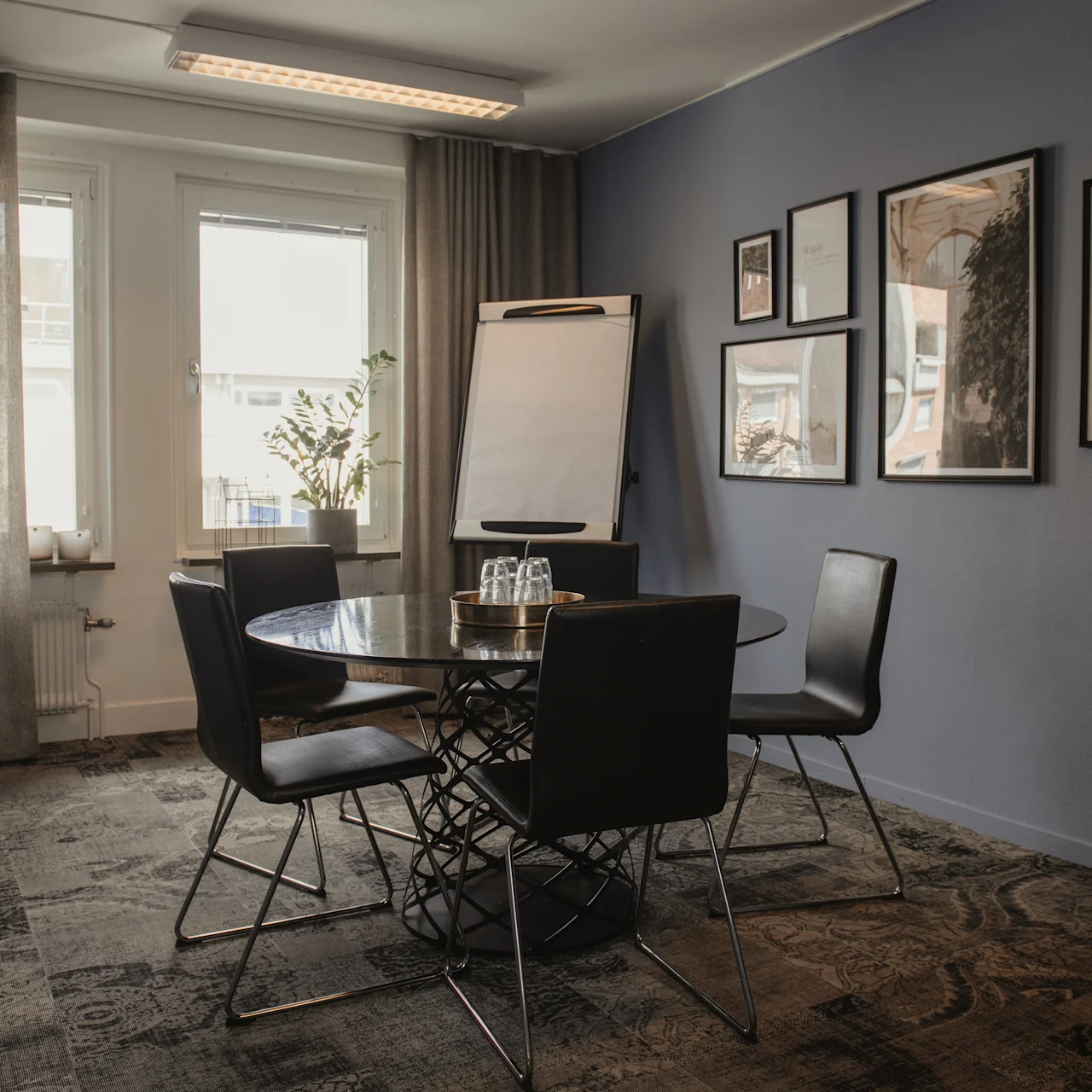 Round table with four chairs in a dimly lit office. A flip chart stands nearby, and framed art hangs on blue walls. Windows with curtains illuminate the gray carpeted floor.