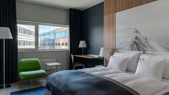 A modern hotel room with a comfortable bed, a bright green armchair, a desk, and a large window overlooking buildings.
