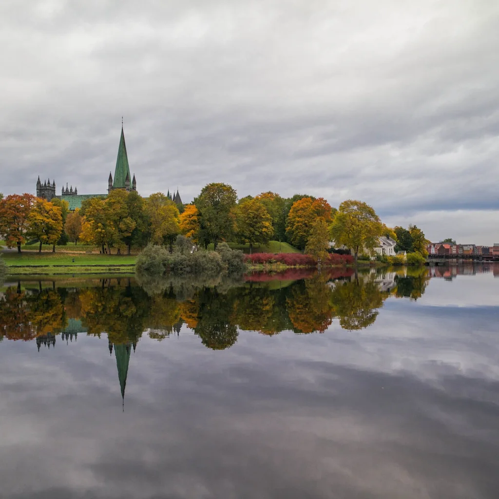 Syksyinen maisema värikkäine puineen ja kirkontorneineen heijastuu tyynestä vedestä pilvisen taivaan alla.