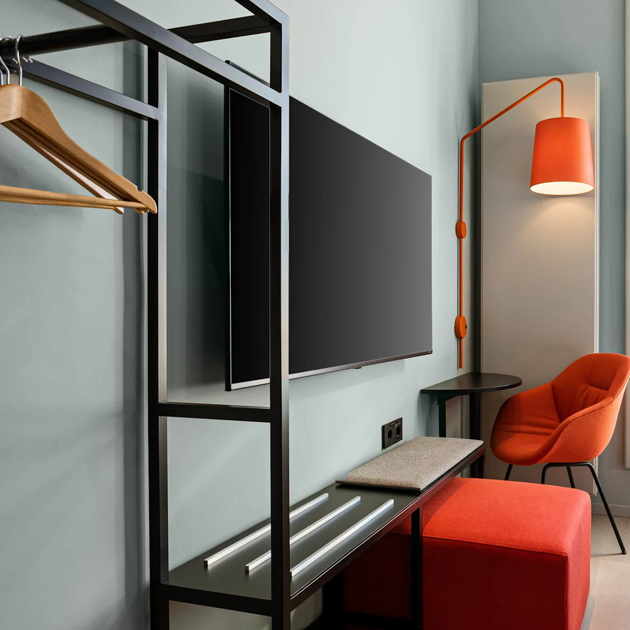 TV mounted on the wall beside a modern stand, with an orange chair under a matching lamp. Large windows light the room, including a bed and minimalist furnishings.