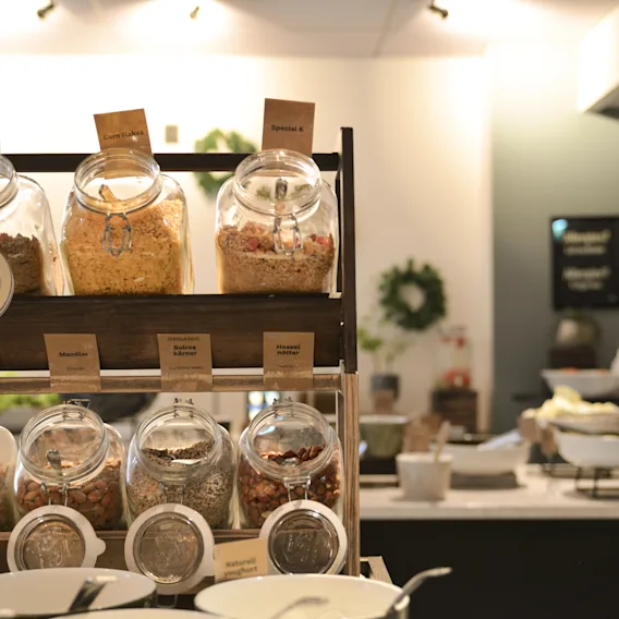 Glass jars filled with various cereals sit on wooden shelves, labeled "ALL BRAN," "Special K," "branflakes," "cornflakes" in a buffet setting. White bowls and serving utensils are in front.