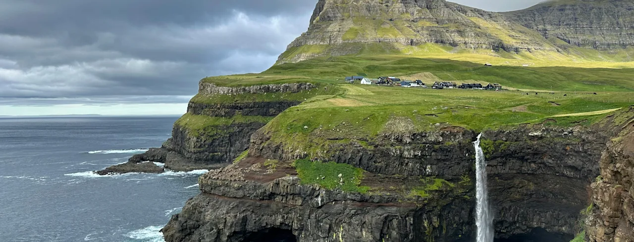 En fantastisk utsikt över ett vattenfall som kastar sig från en grön klippa ner i det mörkblå havet under en molnig himmel. Små hus syns på klipptoppen.