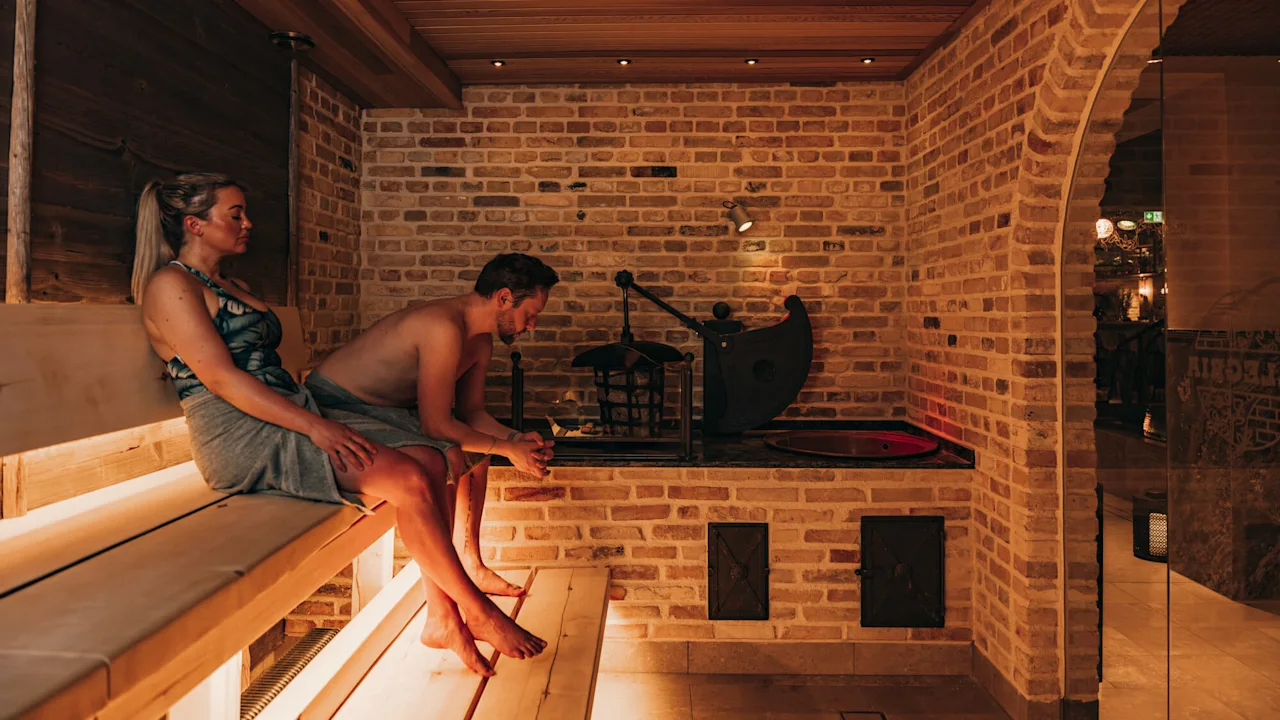 Two people sit on a wooden bench in a sauna, relaxing against a backdrop of warm-toned brick walls. Dim lighting creates a cozy atmosphere.