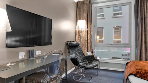 Et moderne hotelværelse med en elegant sort stol og gennemskinnelig skrivebordsstol mod et vægmonteret TV, med store vinduer dækket af gardiner. Teksten inkluderer "BAR & A" udenfor.