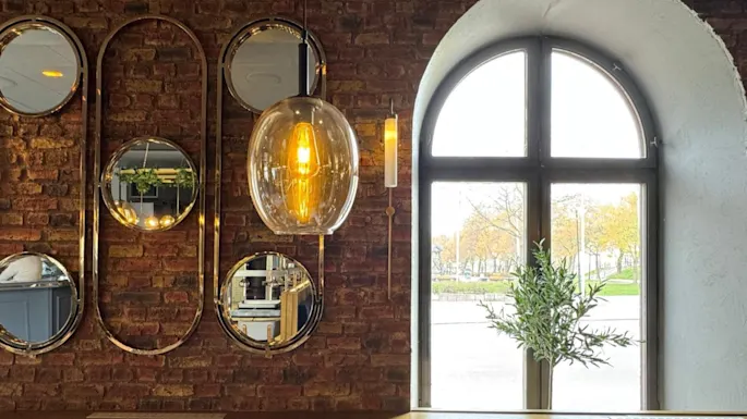 En glødende pendellampe henger nær en murvegg prydet med ovale speil som reflekterer lys. Et stort buet vindu avslører en utendørs utsikt med trær, som forsterker den koselige atmosfæren.