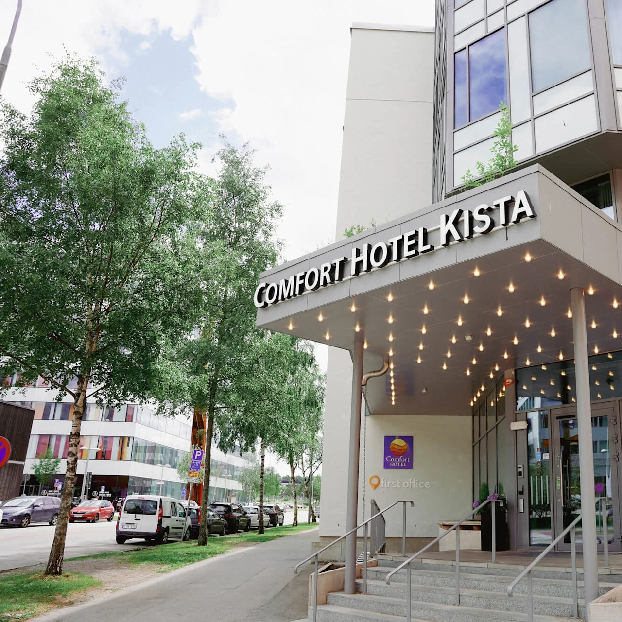 Hotel entrance with a canopy lined with lights, trees on the side, in a city setting.