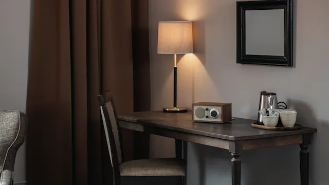 A wooden desk holds a retro radio, lamp, and a tray with a kettle and cups. It's situated near a brown curtain and an empty black-framed mirror on the wall.